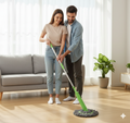A man and a woman in a bright living room, both smiling and looking down at a green and grey mop on a wooden floor. The man is holding the mop's handle, appearing to show the woman how to use it.