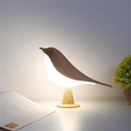 Table Bird Lamp LED on a white desk next to an open book, emitting a warm, soft glow.