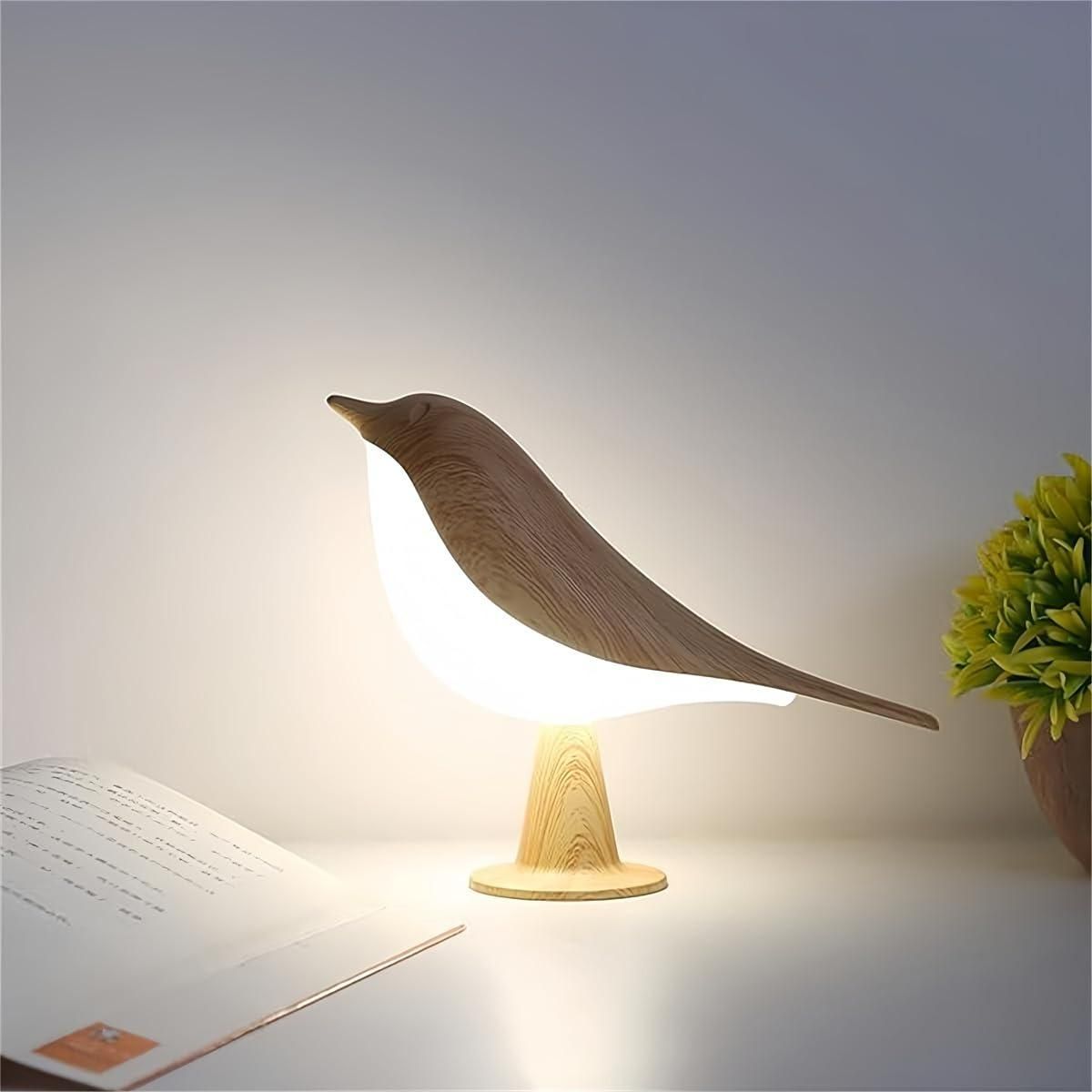Table Bird Lamp LED on a white desk next to an open book, emitting a warm, soft glow.